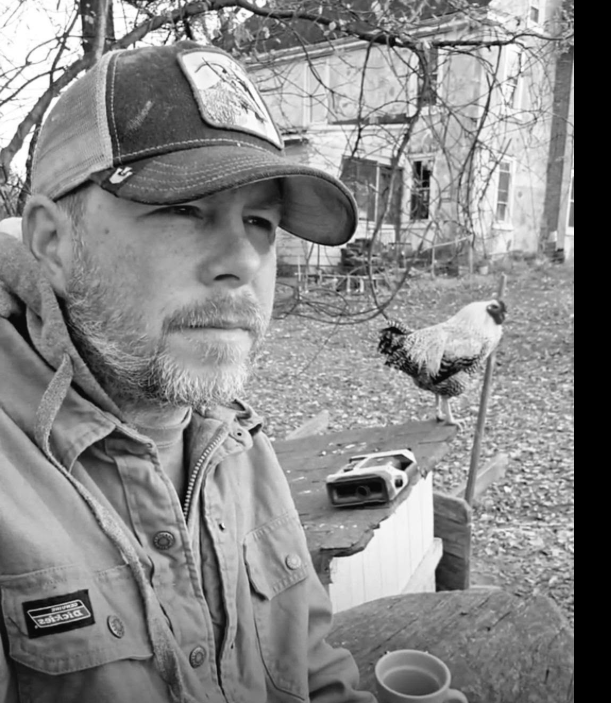 Justin outdoors beside a rooster perched behind him.